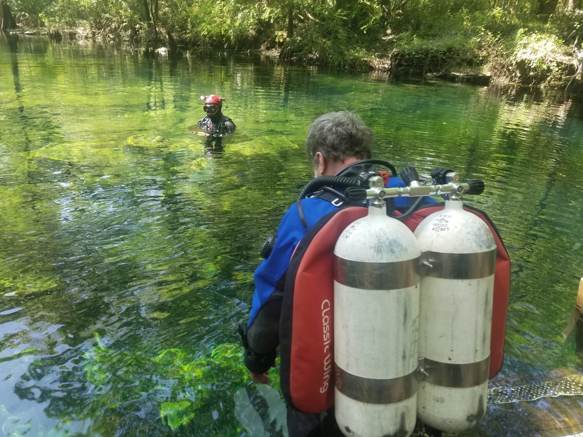 A cave diver in twin tanks wading into a clear spring toward a buddy